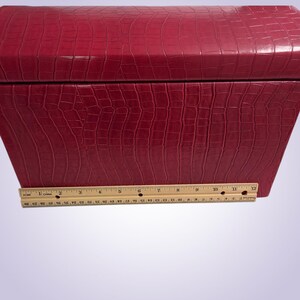 May include: A red, rectangular box with a textured, crocodile-like pattern. The box is closed and has a hinged lid. A wooden ruler is placed at the bottom of the box, measuring approximately 12 inches.