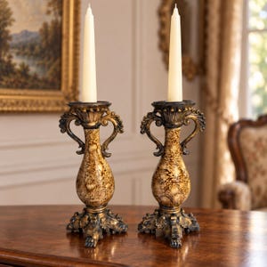 May include: A pair of matching candle holders featuring a textured, mottled gold and black finish with ornate scroll handles and a tripod base. Each holder supports a single, slender, off-white taper candle standing upright in the center.