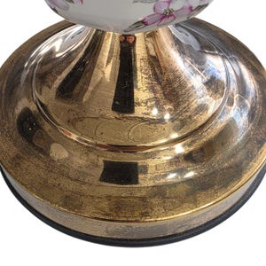 May include: Close-up of a decorative lamp base. The base has a gold metallic finish with a distressed texture. The top portion of the lamp features a white ceramic design with pink floral accents.