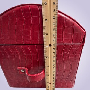 May include: A red, faux-leather jewelry box with a crocodile skin texture. The box is approximately 8 inches tall. It has a handle and a clasp. The box is a deep red color.