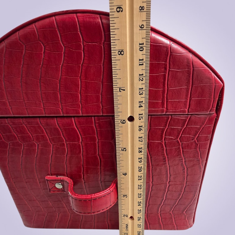 May include: A red, faux-leather jewelry box with a crocodile skin texture. The box is approximately 8 inches tall. It has a handle and a clasp. The box is a deep red color.
