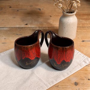 Peut inclure: Deux tasses en céramique avec une conception de poignée unique. Les tasses présentent un dégradé de couleurs, passant d'un dessus brun foncé à une base rouge et noire vibrante. Le texte "Grand Falls" est inscrit sur le devant de chaque tasse.