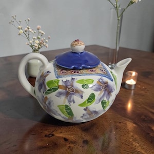 Peut inclure: Une théière en céramique blanche avec un couvercle bleu et un motif floral violet, vert et jaune. La théière a une poignée incurvée et un bec verseur. Le couvercle est surmonté d'un petit bouton décoratif. La théière est posée sur une surface en bois.