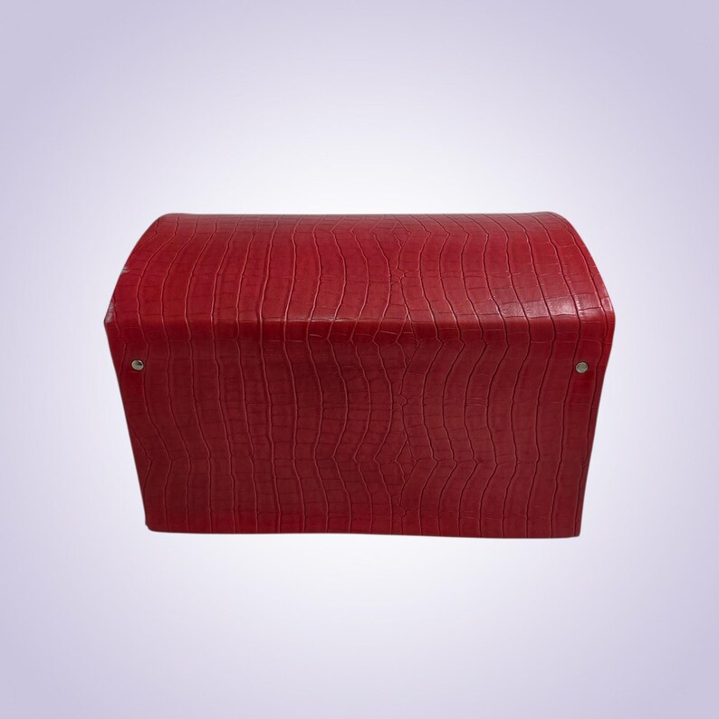 May include: A red, rectangular box with a curved top. The box has a textured, crocodile-like pattern. Two silver rivets are visible on the sides. The box is likely used for storage or display.