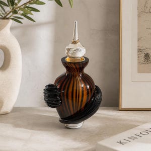 May include: A decorative glass perfume bottle with a clear stopper. The bottle has a brown and black striped pattern with black sculpted accents. The bottle is sitting on a light gray surface.