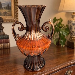 May include: A ceramic vase featuring a glossy finish with a gradient of dark brown at the rim and base, transitioning into a vibrant orange in the center. The vessel has two ornate, curved handles and a fluted, scalloped rim design.