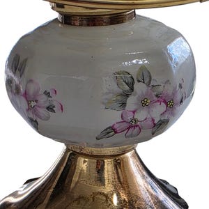 May include: A decorative lamp base with a gold-colored metal base and a white, bulbous body. The body is adorned with hand-painted pink and white floral designs, adding a touch of elegance. The lamp's design is completed with a gold-colored metal top.