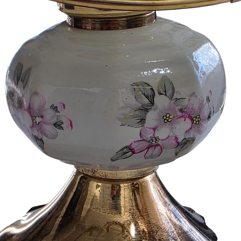 May include: A decorative lamp base with a gold-colored metal base and a white, bulbous body. The body is adorned with hand-painted pink and white floral designs, adding a touch of elegance. The lamp's design is completed with a gold-colored metal top.