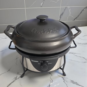Peut inclure: Une mijoteuse Crock-Pot noire avec un couvercle et des poignées. La base est argentée avec un bouton de commande noir et le texte "Versaware". La mijoteuse est posée sur une surface en marbre blanc.