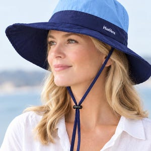 May include: A two-tone sun hat featuring a light blue crown and a wide, dark navy blue brim. The hat includes a dark navy blue chin strap with an adjustable black toggle. The brand name HueVera is printed in white on the front of the brim.