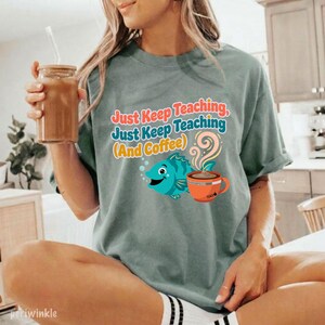 May include: A sage green t-shirt featuring the text "Just Keep Teaching, Just Keep Teaching (And Coffee)" in colorful letters, accompanied by a graphic of a blue fish and an orange coffee mug with steam rising from it.