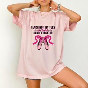 以下が含まれることがあります： 薄いピンク色の半袖Tシャツ。黒とピンクの文字で「TEACHING TINY TOES to Twirl DANCE EDUCATOR」と書かれており、その下にリボンが結ばれたピンクのバレエシューズのグラフィックイラストが描かれています。