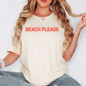 May include: A cream-colored short-sleeve t-shirt featuring the text BEACH PLEASE printed in bold orange capital letters across the chest. The garment has a crew neckline and a relaxed fit, providing a simple and casual appearance for daily wear.
