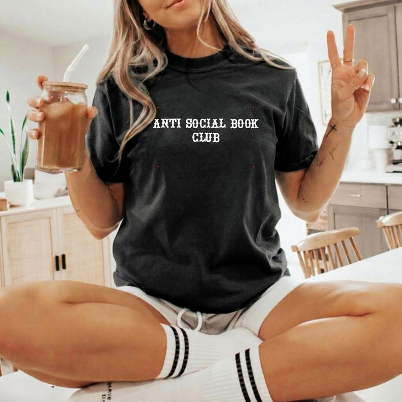 May include: A black short-sleeved t-shirt featuring the white text ANTI SOCIAL BOOK CLUB printed on the chest. The shirt has a crew neckline and a standard fit, paired with white socks that have two black stripes near the top edge.