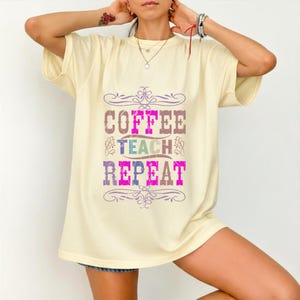 Op de afbeelding: Een lichtgeel T-shirt met korte mouwen en een verweerde grafische print op de voorzijde. De tekst COFFEE TEACH REPEAT staat in gestapelde, veelkleurige blokletters, omlijst door decoratieve paarse krullen aan de boven- en onderkant.