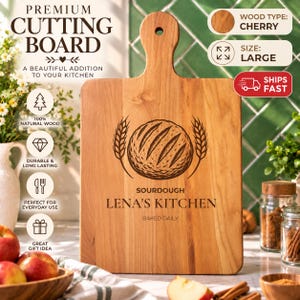 May include: A large cherry wood cutting board with a handle and a hole for hanging. The board features an engraved illustration of a sourdough loaf flanked by wheat stalks, with the text SOURDOUGH LENA'S KITCHEN BAKED DAILY.