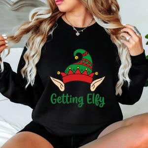 May include: A black long-sleeve sweatshirt featuring a graphic of a green and red elf hat with yellow bells and pointed elf ears. Below the graphic, the text "Getting Elfy" is printed in green script. The garment has a crew neckline.