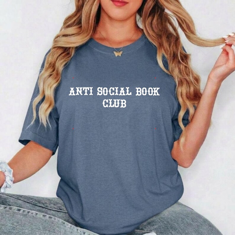 May include: A slate blue short-sleeved t-shirt featuring the white text "ANTI SOCIAL BOOK CLUB" printed in a stylised font across the chest. The garment has a crew neckline and a relaxed fit, providing a casual look for everyday wear.