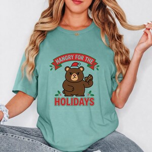 May include: A teal short-sleeve t-shirt featuring a graphic of a brown bear wearing a red Santa hat and holding a bitten gingerbread cookie. The text reads "HANGRY FOR THE HOLIDAYS" in red letters, accented by green holly leaves and red berries.