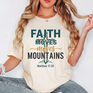 May include: A cream-coloured t-shirt featuring the text "FAITH MOVES MOUNTAINS" in teal and gold. The design includes a cross and mountain graphic, with the Bible verse Matthew 17:20 below. The shirt is a casual, short-sleeved style.