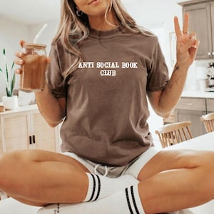May include: A brown short-sleeve t-shirt featuring the white text "ANTI SOCIAL BOOK CLUB" printed on the chest. The shirt has a crew neckline and is worn with white socks featuring two black stripes near the top, paired with white shorts.