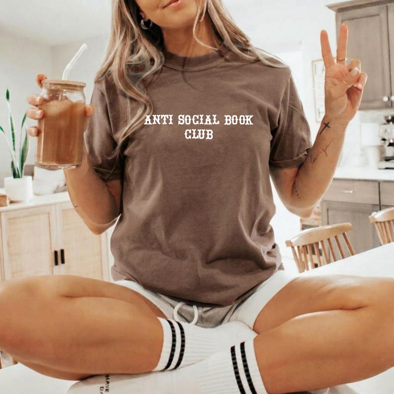 May include: A brown short-sleeve t-shirt featuring the white text "ANTI SOCIAL BOOK CLUB" printed on the chest. The shirt has a crew neckline and is worn with white socks featuring two black stripes near the top, paired with white shorts.
