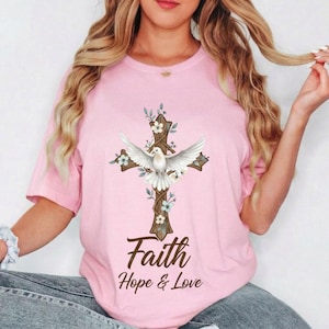 May include: Pale pink t-shirt with a cross design, a white dove, and floral details. The cross is brown, and the dove has open wings. The text "Faith Hope & Love" is printed below the cross in a brown script font.