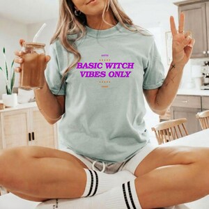 Pode incluir: Uma t-shirt de manga curta verde-clara com o texto roxo "BASIC WITCH VIBES ONLY" estampado no peito. A t-shirt tem gola redonda e um corte descontraído, combinada com meias brancas com riscas pretas na extremidade superior.