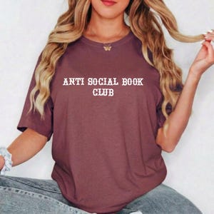 May include: A maroon short-sleeved t-shirt featuring the white text "ANTI SOCIAL BOOK CLUB" printed in a bold, serif font across the chest. The garment has a classic crew neckline and a relaxed fit, providing a simple and straightforward design.
