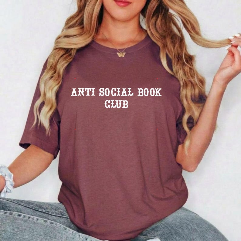 May include: A maroon short-sleeved t-shirt featuring the white text "ANTI SOCIAL BOOK CLUB" printed in a bold, serif font across the chest. The garment has a classic crew neckline and a relaxed fit, providing a simple and straightforward design.