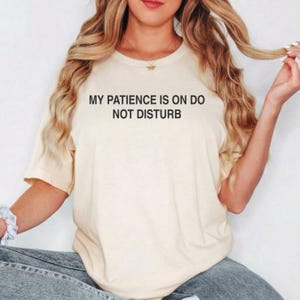 May include: A cream-coloured short-sleeve t-shirt featuring the black text MY PATIENCE IS ON DO NOT DISTURB printed across the chest. The garment has a crew neckline and a relaxed fit, providing a simple and straightforward design for casual wear.