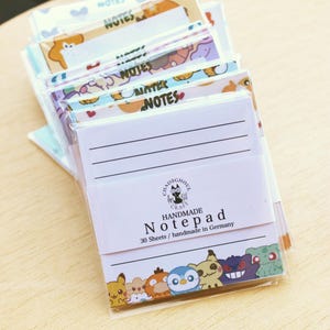 Könnte beinhalten: Ein Stapel handgefertigter Notizblöcke in transparenter Kunststoffverpackung. Jeder Notizblock hat liniertes Papier mit bunten Cartoon-Figuren am unteren Rand. Das Etikett trägt die Aufschrift Chaosghoul Craft Handmade Notepad 30 Sheets / handmade in Germany.