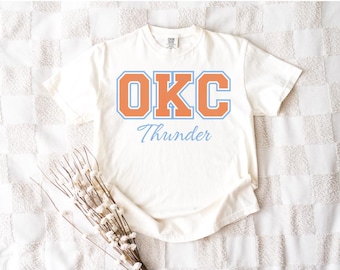 May include: Cream-colored t-shirt featuring the text "OKC Thunder" in orange and light blue. The word "OKC" is in a bold, collegiate-style font, with "Thunder" in a script font below. The shirt is displayed on a white, textured surface.