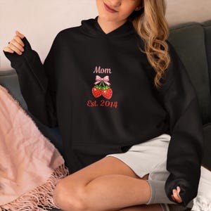 Puede incluir: Sudadera negra con lazo rosa y diseño de fresas. El texto "Mom Est. 2014" está impreso en rosa. Una prenda informal para el uso diario.