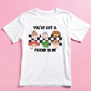 May include: A white short-sleeve t-shirt featuring a black and white checkered pattern with cartoon character faces including a horse, space ranger, dog, cowgirl, dinosaur, and cowboy. The text reads YOU'VE GOT A FRIEND IN ME in black letters.