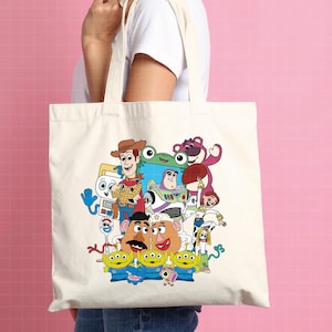 May include: A cream-coloured tote bag featuring a colourful graphic of characters from the animated film Toy Story. The design includes Woody, Buzz Lightyear, Mr. Potato Head, and the Little Green Men. The bag has two cream-coloured handles.