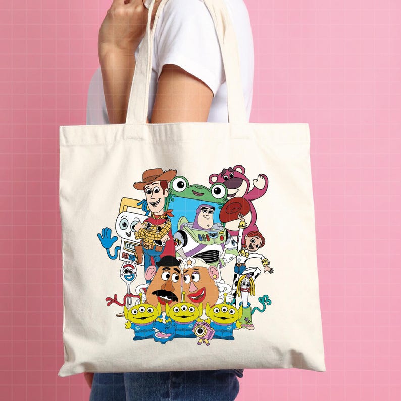 May include: A cream-coloured tote bag featuring a colourful graphic of characters from the animated film Toy Story. The design includes Woody, Buzz Lightyear, Mr. Potato Head, and the Little Green Men. The bag has two cream-coloured handles.