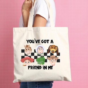 May include: A cream-colored canvas tote bag featuring a black and white checkered pattern background with cartoon character faces including a horse, space ranger, dog, cowgirl, dinosaur, and cowboy, with the text YOU'VE GOT A FRIEND IN ME.