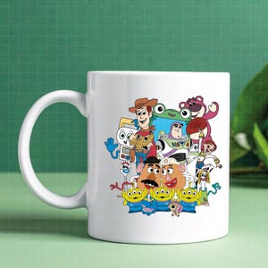 May include: White ceramic mug with a colourful graphic of characters from the animated film Toy Story. The design includes Woody, Buzz Lightyear, Mr. Potato Head, and other recognisable characters. The mug has a curved handle.