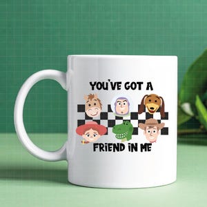 May include: A white ceramic mug featuring a black and white checkered pattern with six cartoon character faces including a horse, space ranger, dog, cowgirl, dinosaur, and cowboy, with the text "YOU'VE GOT A FRIEND IN ME" printed in black.