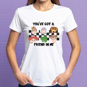 May include: A white short-sleeved t-shirt featuring a black and white checkered pattern with cartoon character faces including a horse, space ranger, dog, cowgirl, dinosaur, and cowboy. The text reads YOU'VE GOT A FRIEND IN ME in black letters.