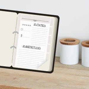 May include: A black ring-bound recipe book with white pages featuring the German text ZUTATEN and ZUBEREITUNG, positioned next to a rectangular black cast iron skillet on a wooden base containing cooked meat and green vegetables.