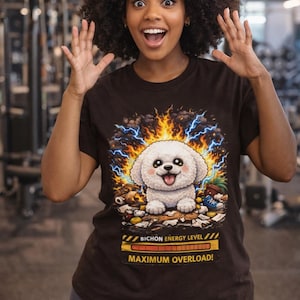 Puede incluir: Camiseta marrón con un diseño de perro Bichon Frise de dibujos animados, rodeado de rayos y llamas. El texto dice "Bichon Energy Level - Maximum Overload!". La persona que lleva la camiseta sonríe y tiene los brazos levantados.