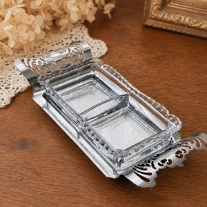 May include: A rectangular silver-toned metal serving tray with ornate, cut-out handles on each end. The tray holds a clear, two-compartment glass dish with a beaded rim, designed for serving small portions of food or condiments on a table.