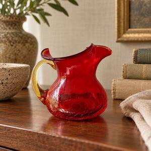 May include: A small, vibrant red glass pitcher featuring a distinct crackle texture throughout its rounded body. The vessel includes a curved, translucent amber-colored handle and a wide, flared pouring spout, showcasing a classic design.
