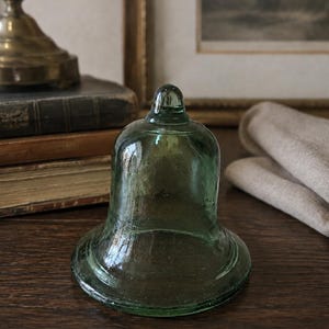 May include: A bell-shaped object made of translucent, light green glass. The item features a smooth, curved exterior with a small, rounded knob at the top and a wide, flared base. The glass has a subtle, slightly uneven texture throughout its surface.