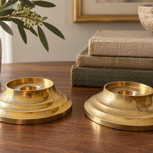 May include: Two circular brass candle holders featuring a stepped, tiered base design with a central opening for a candle. The metal surface has a polished, reflective finish with subtle variations in tone and texture across the round forms.
