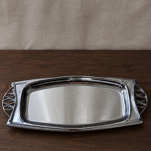 May include: A rectangular silver-toned metal serving tray with a polished finish. The tray features a smooth, flat central surface and raised edges. On each side, there are decorative handles with a cutout design consisting of three oval shapes.