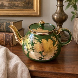 May include: A round, dark green ceramic teapot featuring a gold-colored chinoiserie landscape pattern. The teapot has a matching lid with a gold-colored finial, a gold-colored spout, and a gold-colored handle. The surface is glossy and smooth.