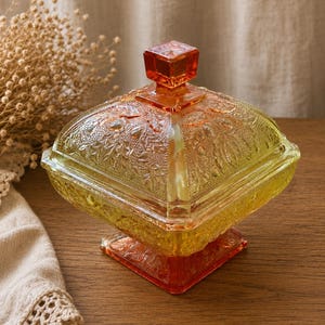 May include: A square glass candy dish with a lid, featuring a gradient color transition from yellow to orange. The glass surface is decorated with an embossed floral and leaf pattern, and the lid is topped with a small, square-shaped handle.